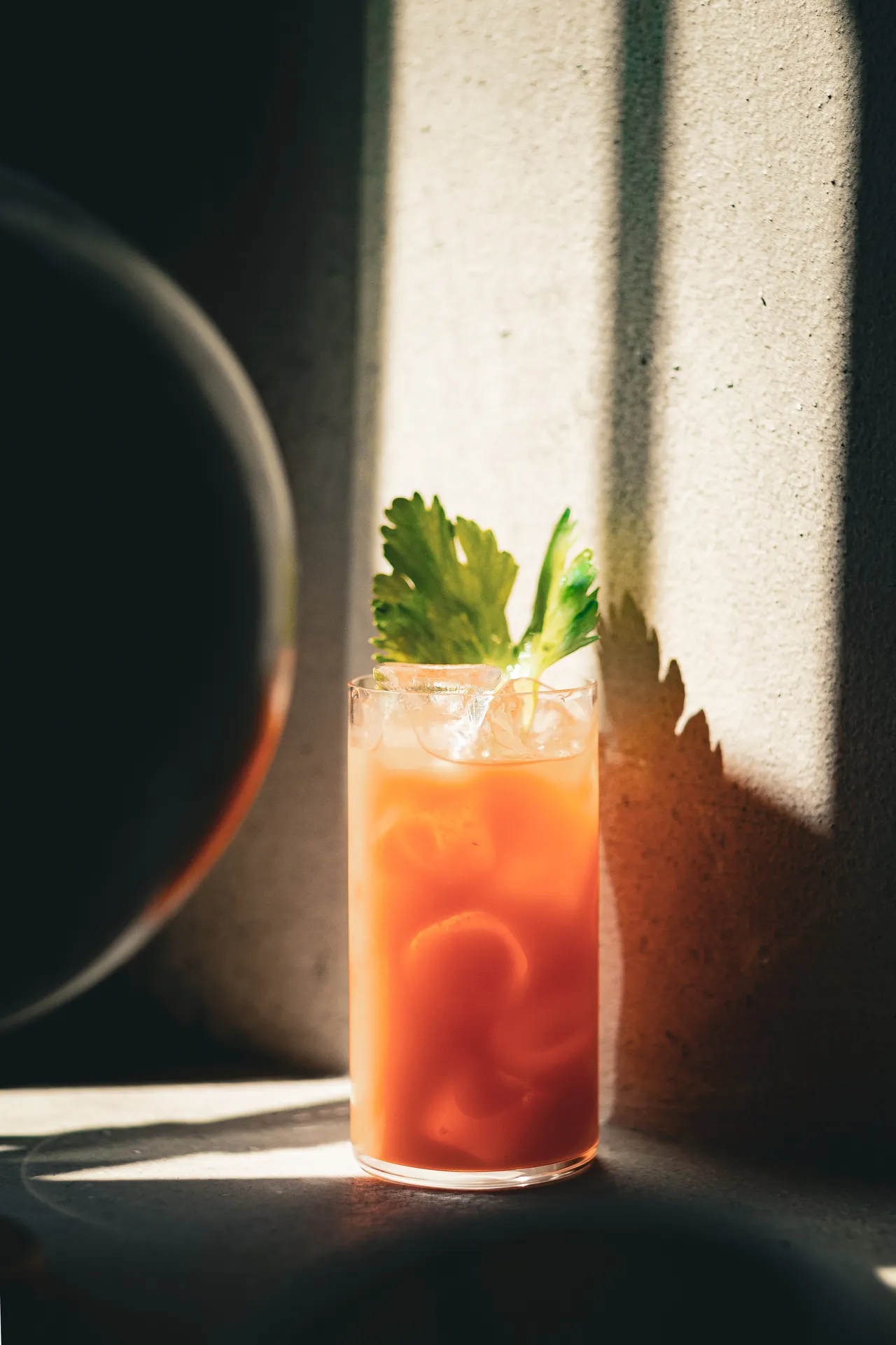 Orange highball garnished with celery in a blade of light, beside a glass sphere