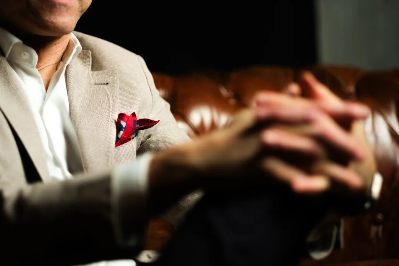 Close-up of clasped hands, a pale jacket with a red pocket square against a leather Chesterfield sofa