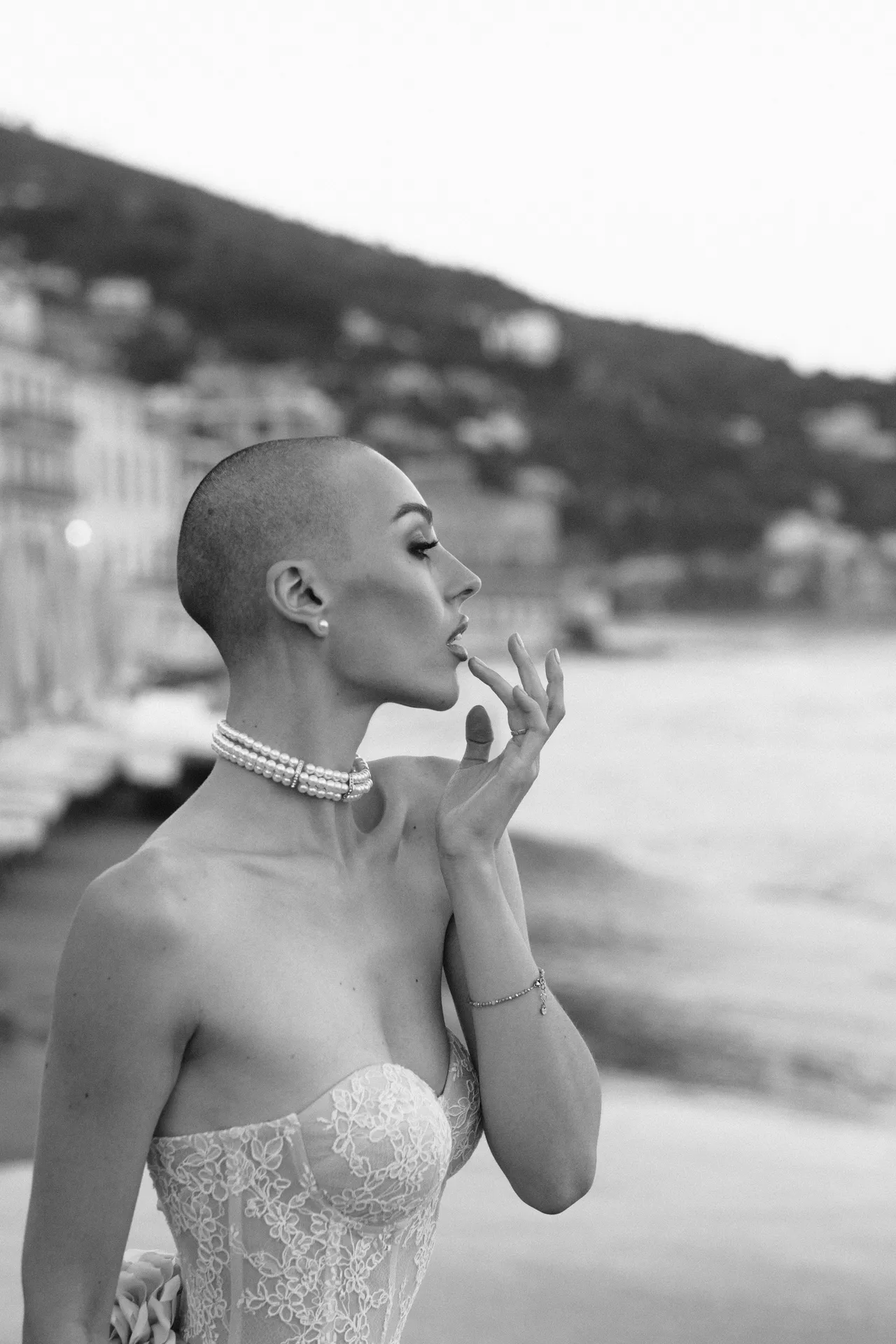 Profile of a woman wearing a pearl choker and a lace dress, the coastline blurred behind her
