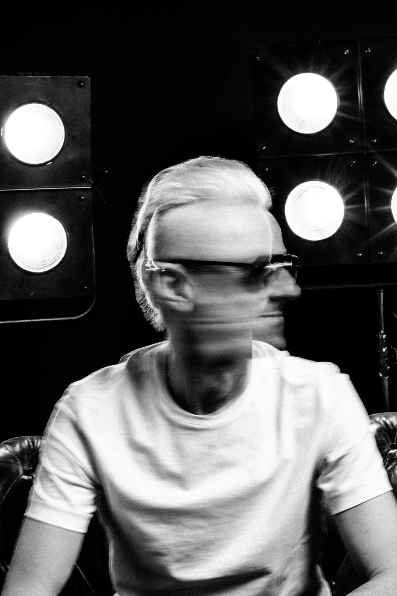 Black and white portrait of a man in a white T-shirt, his face blurred by motion in front of a wall of lit bulbs
