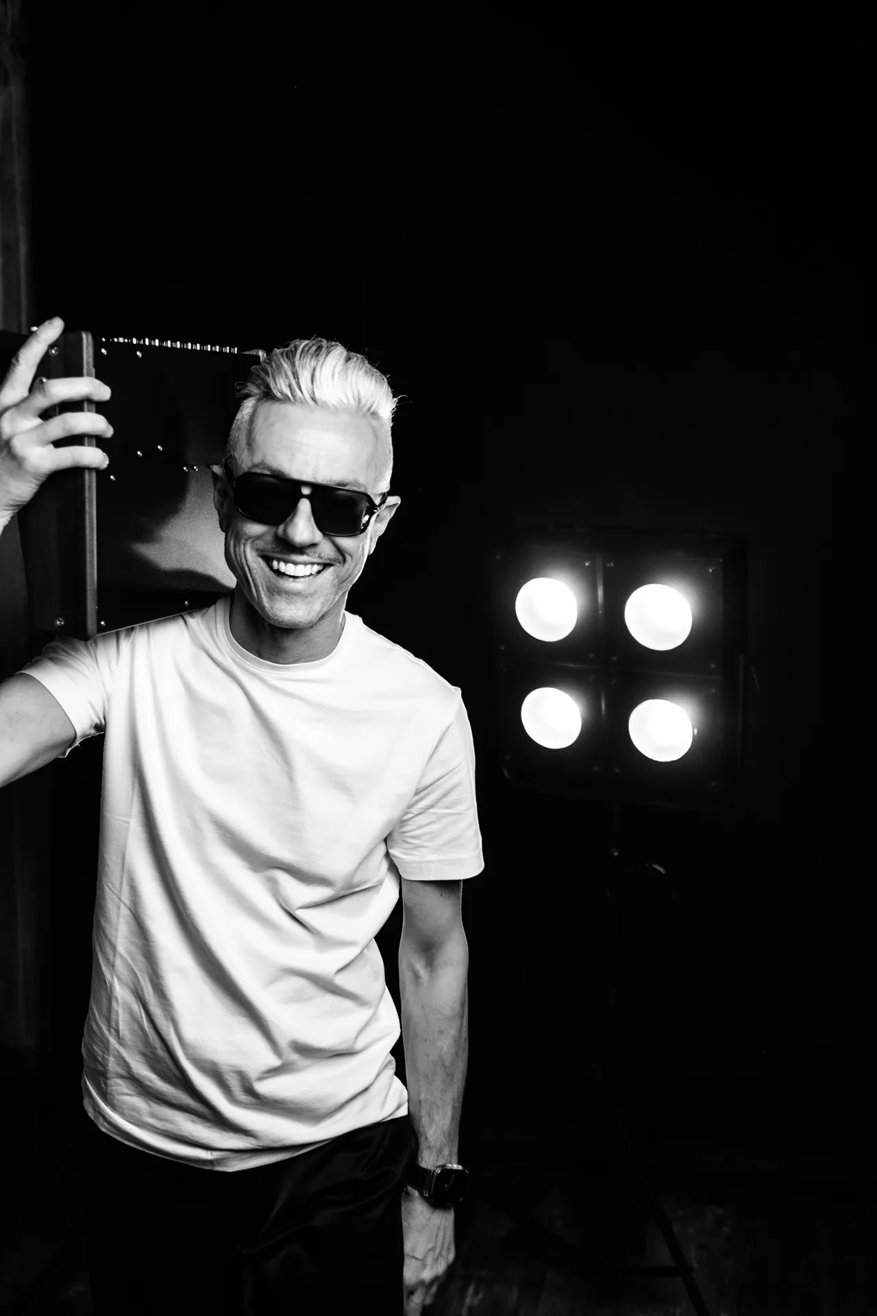 Smiling man with dark glasses and platinum hair beside four glowing bulbs, in black and white