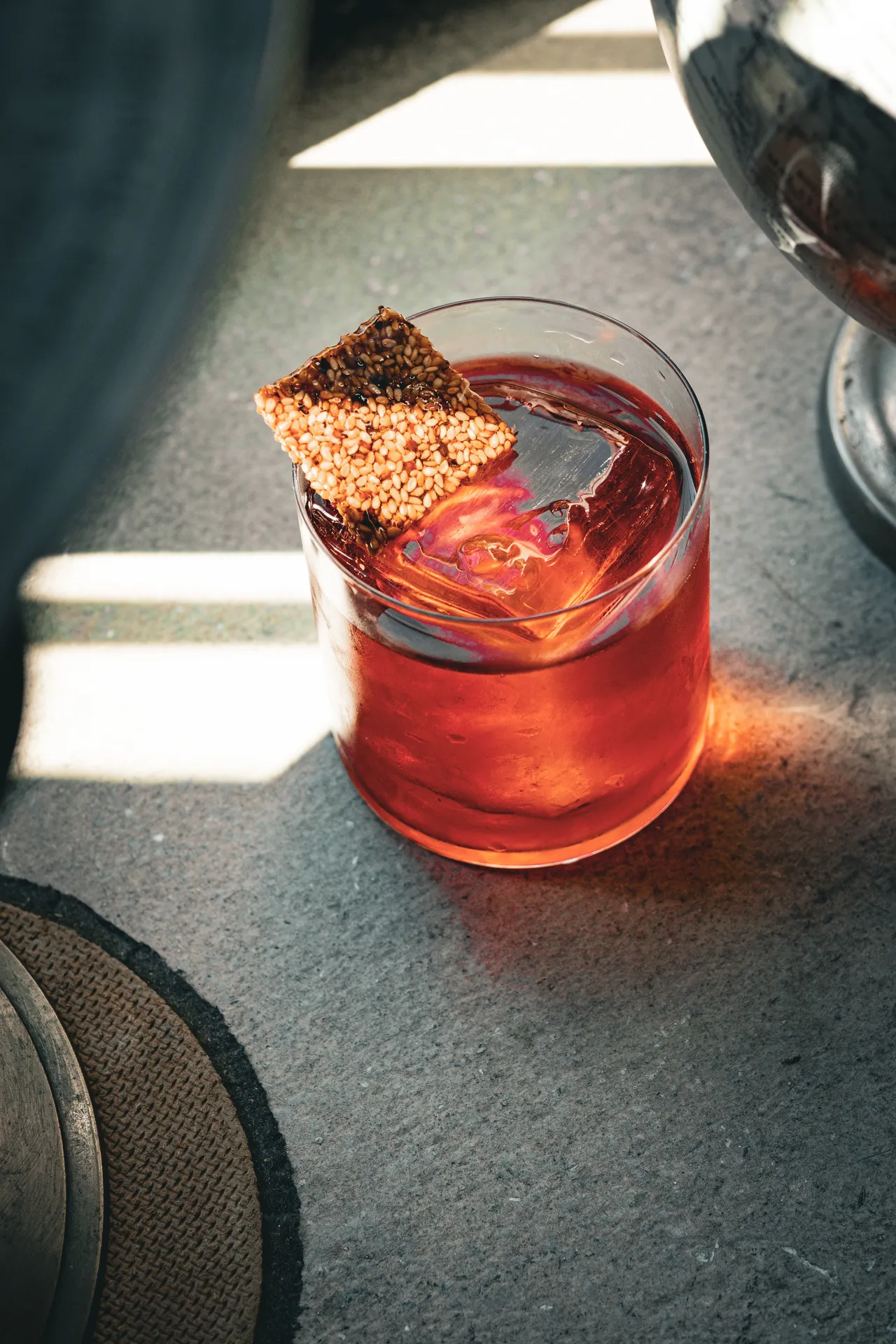 Tumbler of red cocktail topped with a crisp wafer, window light on concrete