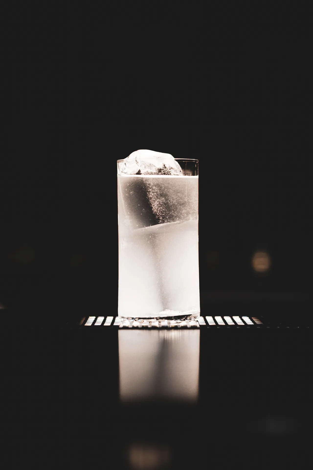 Collins glass with ice on a striped coaster, reflected on a glossy surface in the dark